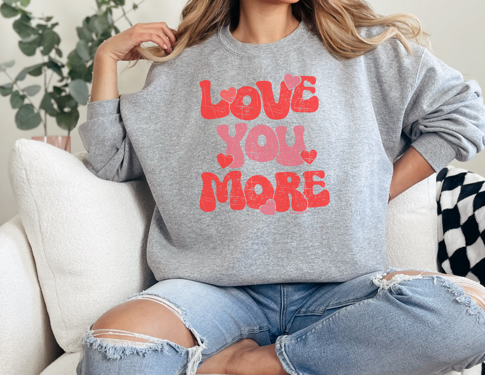 A person in jeans sitting comfortably on a chair, showcasing the Love You More Crew sweatshirt by Worlds Worst Tees. Unisex, cotton-polyester blend, loose fit, ribbed knit collar, no itchy seams.