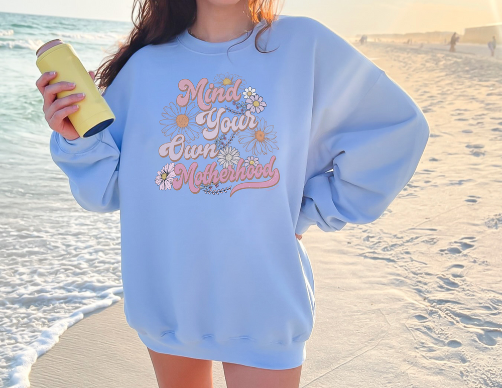A woman at the beach holds a can, showcasing the Mind Your Own Motherhood Crew sweatshirt. Unisex, heavy blend fabric, ribbed knit collar, no itchy seams, loose fit. Ideal comfort for all.