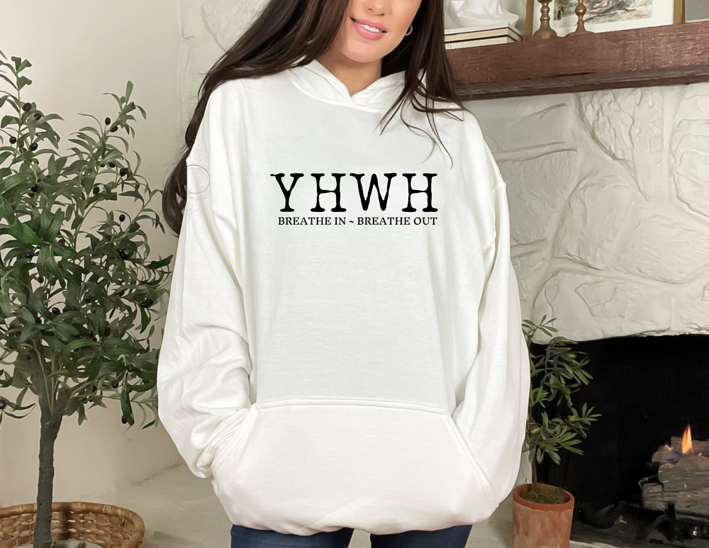 A woman in a white YHWH Hoodie, posing next to a houseplant. Unisex heavy blend hooded sweatshirt, cotton-polyester mix, plush and warm, kangaroo pocket, drawstring hood. Perfect for cold days.