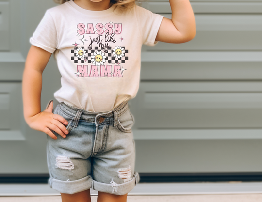 A girl in a white shirt and shorts poses wearing the Sassy Like My Mama Kids Tee from Worlds Worst Tees. Made of soft 100% cotton, perfect for active kids. Classic fit for all-day comfort.