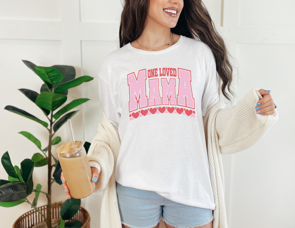 A woman in a casual dress holding a drink, showcasing the One Loved Mama Tee from Worlds Worst Tees. Unisex, heavy cotton tee with ribbed knit collar, tape shoulders, and classic fit. No side seams for comfort.