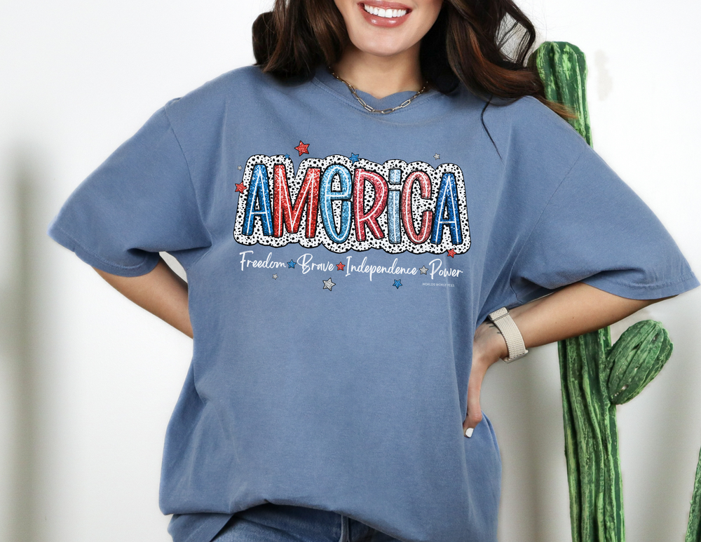 A woman in a blue America Tee, showcasing a relaxed fit and ring-spun cotton fabric. Garment-dyed for extra coziness, with double-needle stitching for durability and a seamless design.
