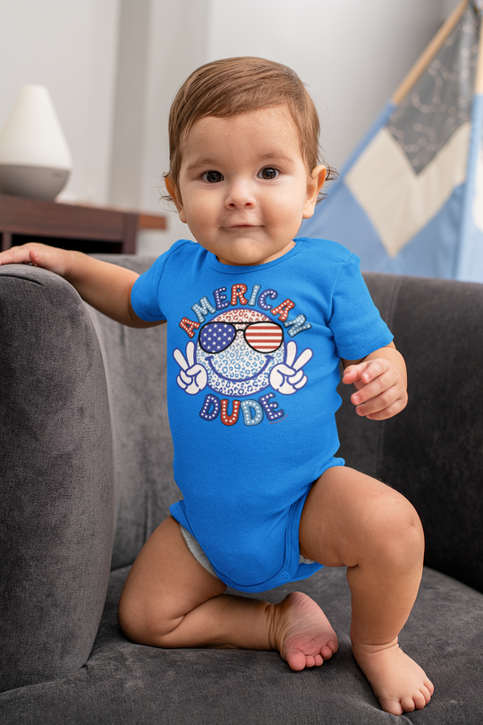 An American Dude Onesie for infants, featuring a blue shirt with a cartoon face and sunglasses. Made of 100% cotton, with ribbed bindings and plastic snaps for easy changing. Ideal for toddlers.