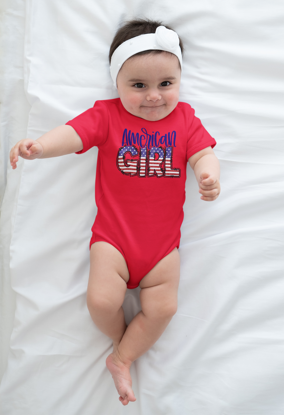 A baby lies on a white blanket, wearing an American Girl Onesie. Close-up of baby's head, foot, and hand. Infant fine jersey bodysuit, 100% cotton, ribbed knitting for durability, plastic snaps for easy changing.