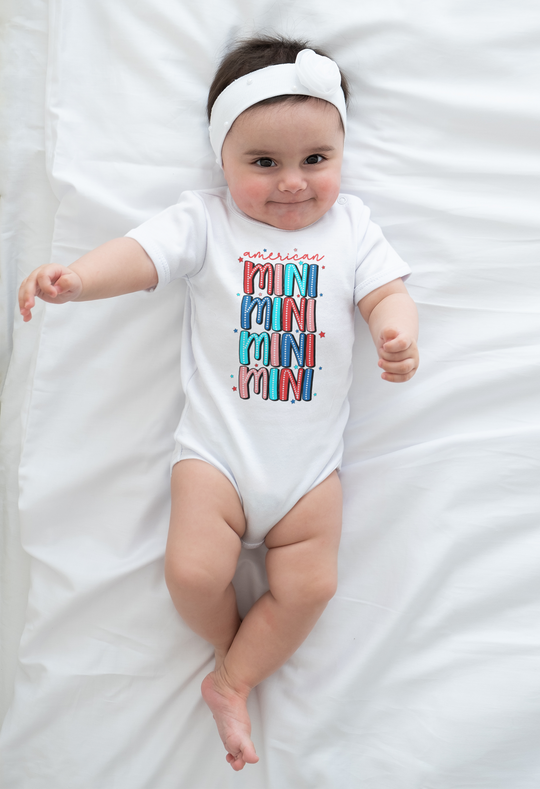 An American Mini Onesie for infants, featuring a baby lying on a white blanket. Made of 100% cotton, with ribbed knitting for durability and plastic snaps for easy changing access. Ideal for newborns to 24M.