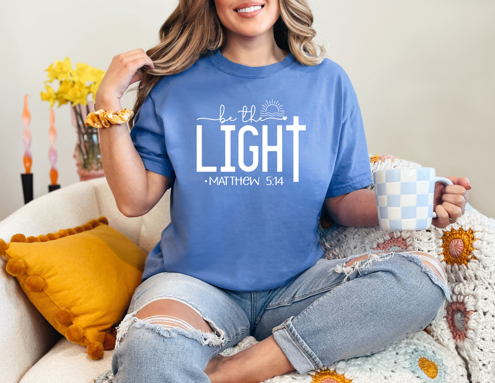 A woman in a blue shirt sitting on a couch, holding a mug. Be the Light Tee: 100% ring-spun cotton, garment-dyed for coziness, with double-needle stitching for durability and a relaxed fit.
