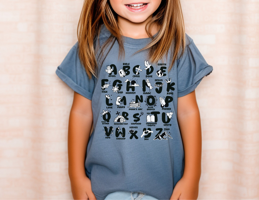 A girl in a Bible Alphabet Kids Tee, smiling, wearing a blue shirt with letters. Made of 100% combed ringspun cotton for comfort and agility. Classic fit, perfect for active kids.