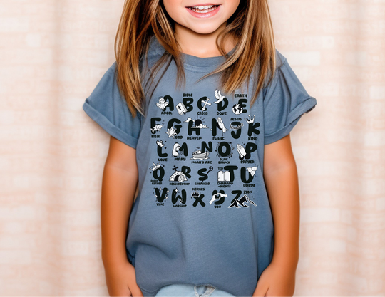 A girl in a Bible Alphabet Kids Tee, smiling, wearing a blue shirt with letters. Made of 100% combed ringspun cotton for comfort and agility. Classic fit, perfect for active kids.