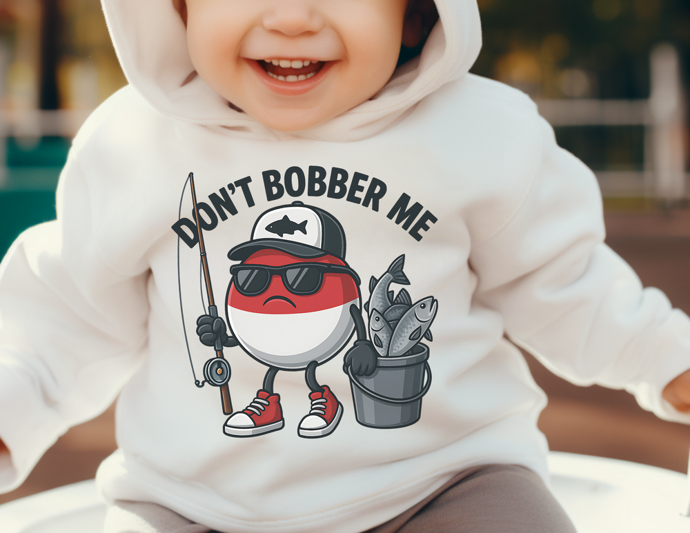 Child wearing Don't Bobber Me Toddler Hoodie featuring a cartoon bobber with fishing gear. The hoodie offers cozy comfort with jersey-lined hood and side-seam pockets.