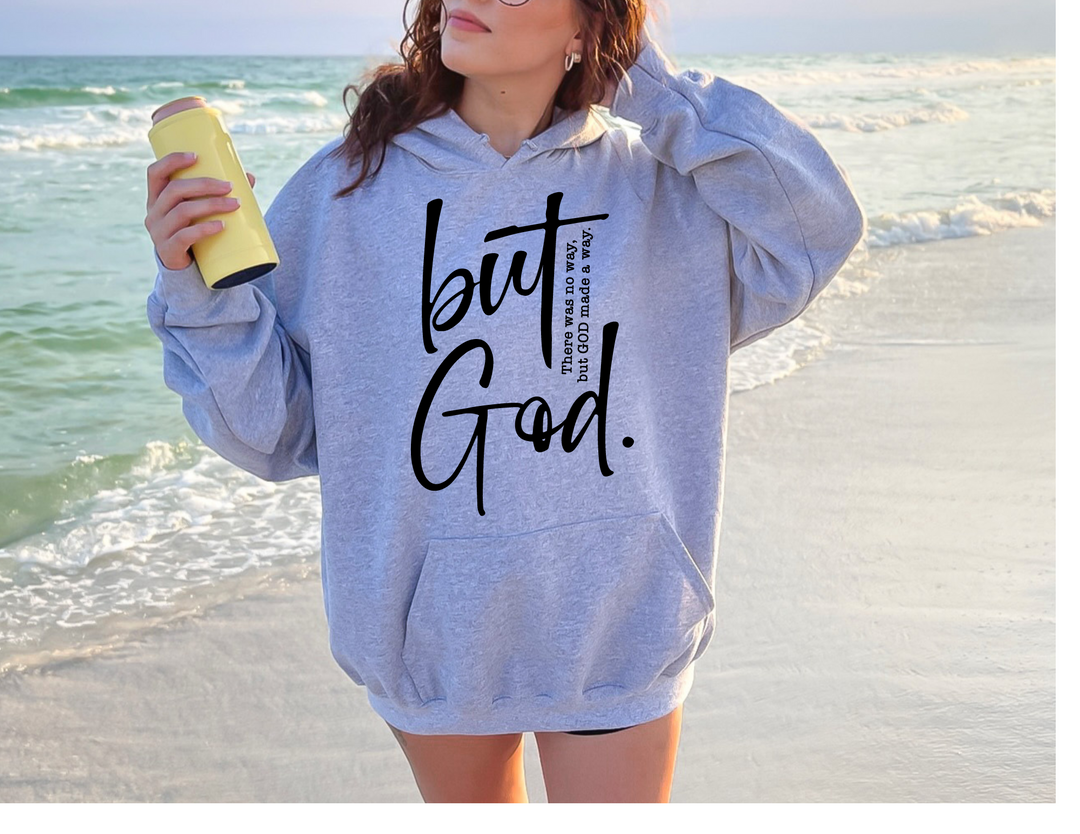 A woman in a grey sweatshirt holds a can on the beach, showcasing the But God Hoodie from Worlds Worst Tees. Unisex heavy blend, cotton-polyester fabric, kangaroo pocket, and matching drawstring.