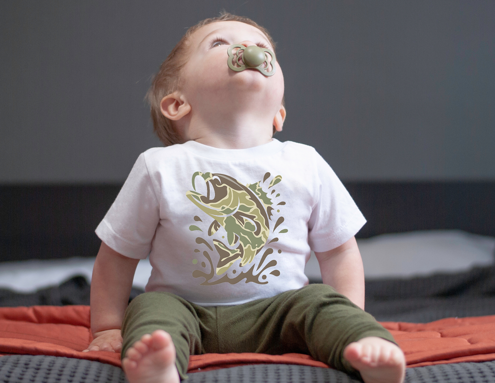 Baby wearing a Camo Fish Baby Tee sits on a bed, featuring a stylized jumping fish design. The white tee is soft, durable, and shrink-resistant.