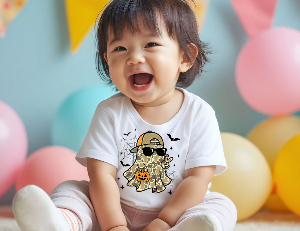 Toddler wearing Candy Ghost Baby Tee with a camo ghost, sunglasses, and jack-o’-lantern design, surrounded by pastel balloons and party decor.