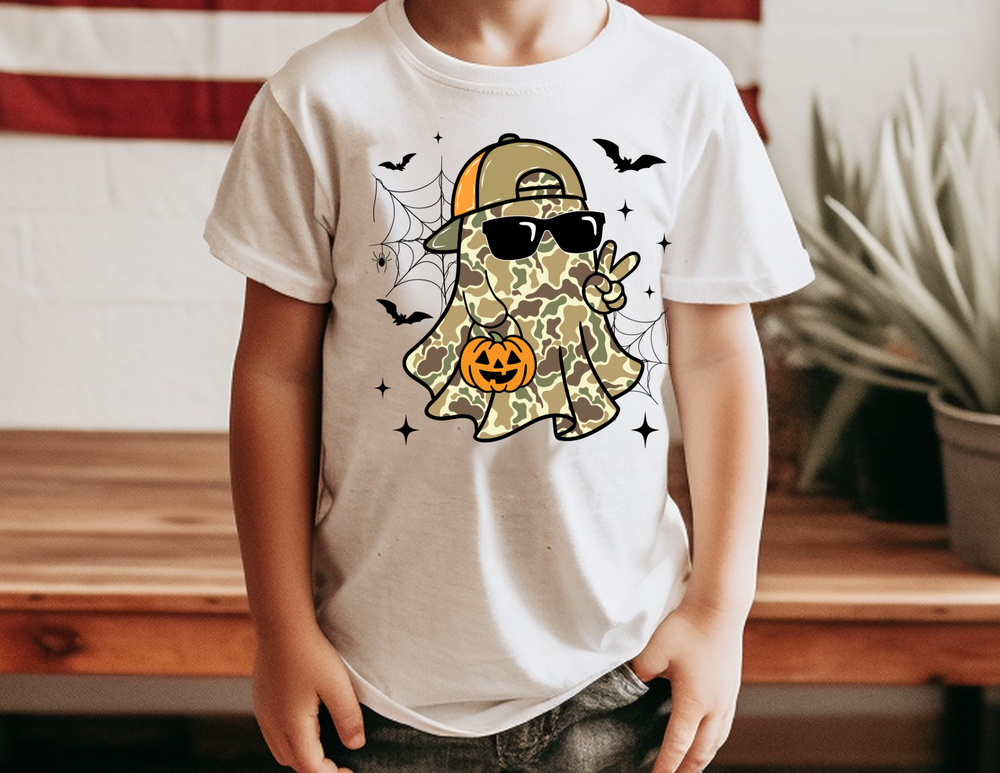Toddler wearing Candy Ghost Toddler Tee featuring a camouflage-patterned ghost with sunglasses, peace sign, and jack-o’-lantern, surrounded by bats, spiderwebs, and sparkles.