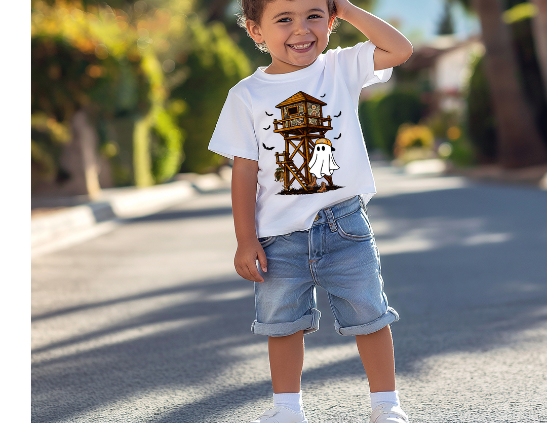 Ghost Hunter Toddler Tee features a Halloween-themed graphic with a tower, ghost, and bats. Worn by a smiling boy on a sunny street.