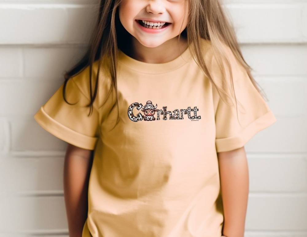 A smiling girl in a Cowhartt Cow Toddler Tee, featuring soft, durable fabric perfect for sensitive skin. Classic fit with a high-quality print for little adventurers. Sizes: 2T, 3T, 4T, 5-6T.
