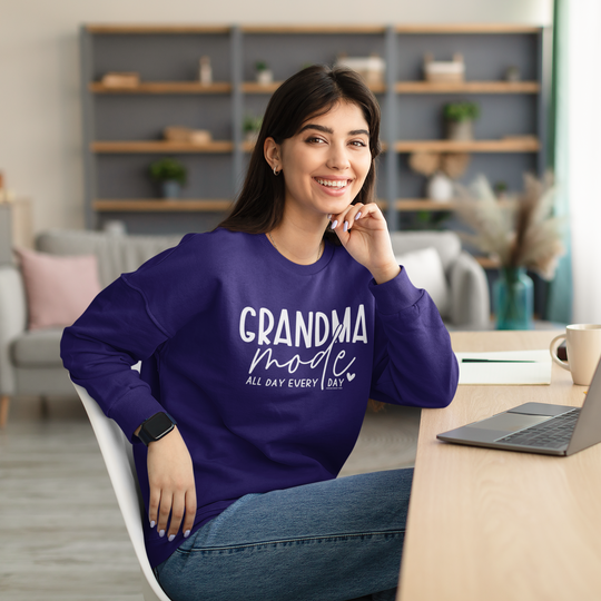 A woman in Grandma Mode Crew sweatshirt, sitting at a table with a laptop, embodying comfort and style. Unisex heavy blend, loose fit, ribbed knit collar, 50% cotton, 50% polyester.
