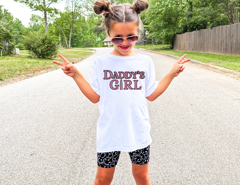 Child wearing Daddy's Girl Kids Tee, white shirt, outdoors. The tee offers comfort from 100% combed ring-spun cotton, perfect for active kids.