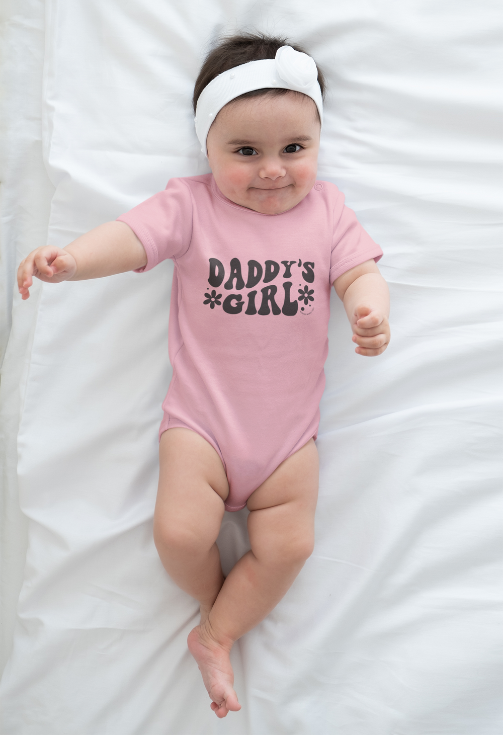 A baby lies on a white surface, wearing a Daddy's Girl Onesie. Close-ups show the infant's head, hand, foot, and nose. This fine jersey bodysuit is 100% cotton, featuring ribbed knitting for durability and plastic snaps for easy changing access.
