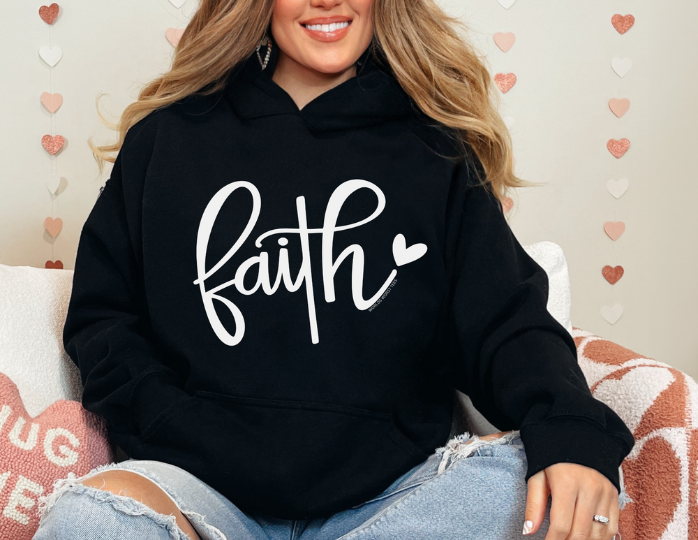 A unisex Faith Hoodie in black, showcasing a woman in a cozy sweatshirt on a couch. Made of cotton and polyester, it's warm and soft with a kangaroo pocket and matching drawstring hood.