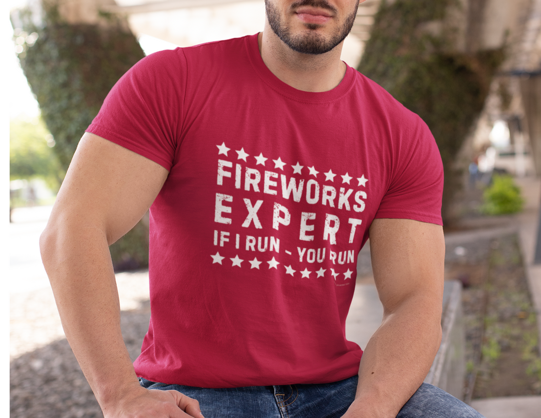 A man in a red Firework Expert Tee, sitting on a bench. Quality cotton tee with ribbed knit collar, taping on shoulders, and dual side seams for shape retention. From Worlds Worst Tees.