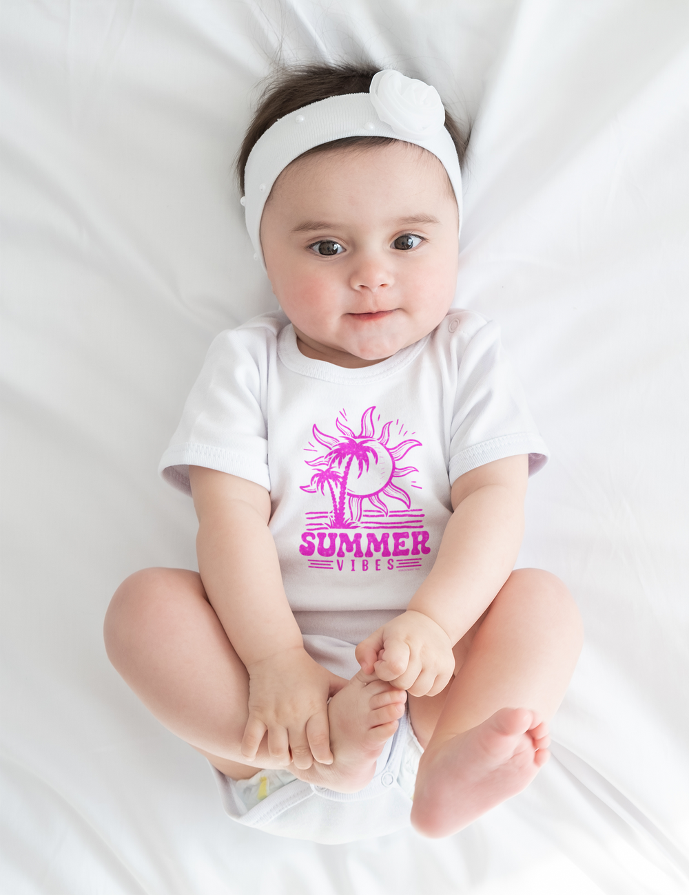 A baby lies on a white sheet wearing a Summer Vibes Onesie. This infant fine jersey bodysuit is 100% cotton, featuring ribbed bindings and plastic snaps for easy changes. Perfect for little ones enjoying the sun and palm trees.