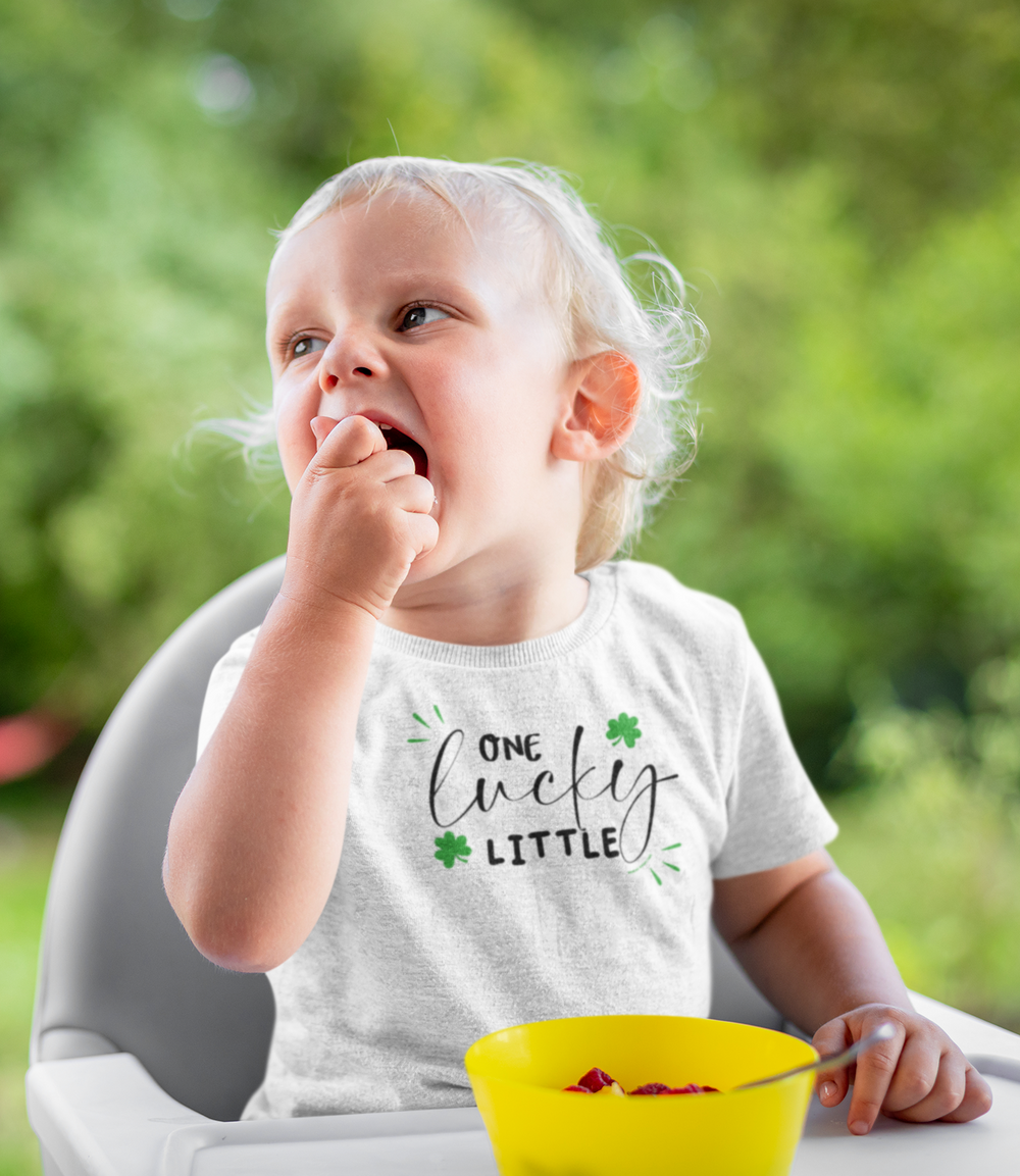 A toddler in a white tee with One Lucky Little Tee print, sitting in a chair, eating fruit. Made of soft 100% combed ringspun cotton, tear-away label, classic fit. Sizes: 2T, 3T, 4T, 5-6T.