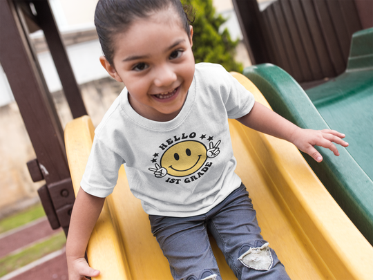 A smiling girl in a Hello 1st Grade Kids Tee, featuring 100% cotton fabric, twill tape shoulders, and ribbed collar. Classic fit, tear-away label, no side seams. Perfect for everyday wear.