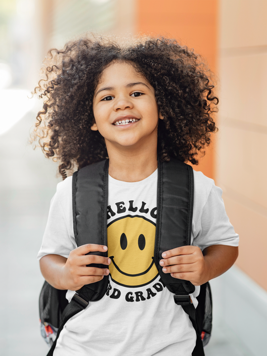 A young girl with curly hair wearing a backpack, showcasing the Hello 3rd Grade Kids Tee from Worlds Worst Tees. 100% cotton, light fabric, classic fit, tear-away label, and durable twill tape shoulders.