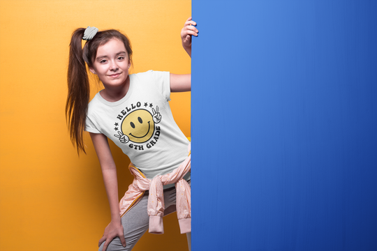 A girl in a Hello 6th Grade Kids Tee, posing with a yellow smiley face on a white shirt. 100% cotton, light fabric, tear-away label, classic fit. Ideal for everyday wear.