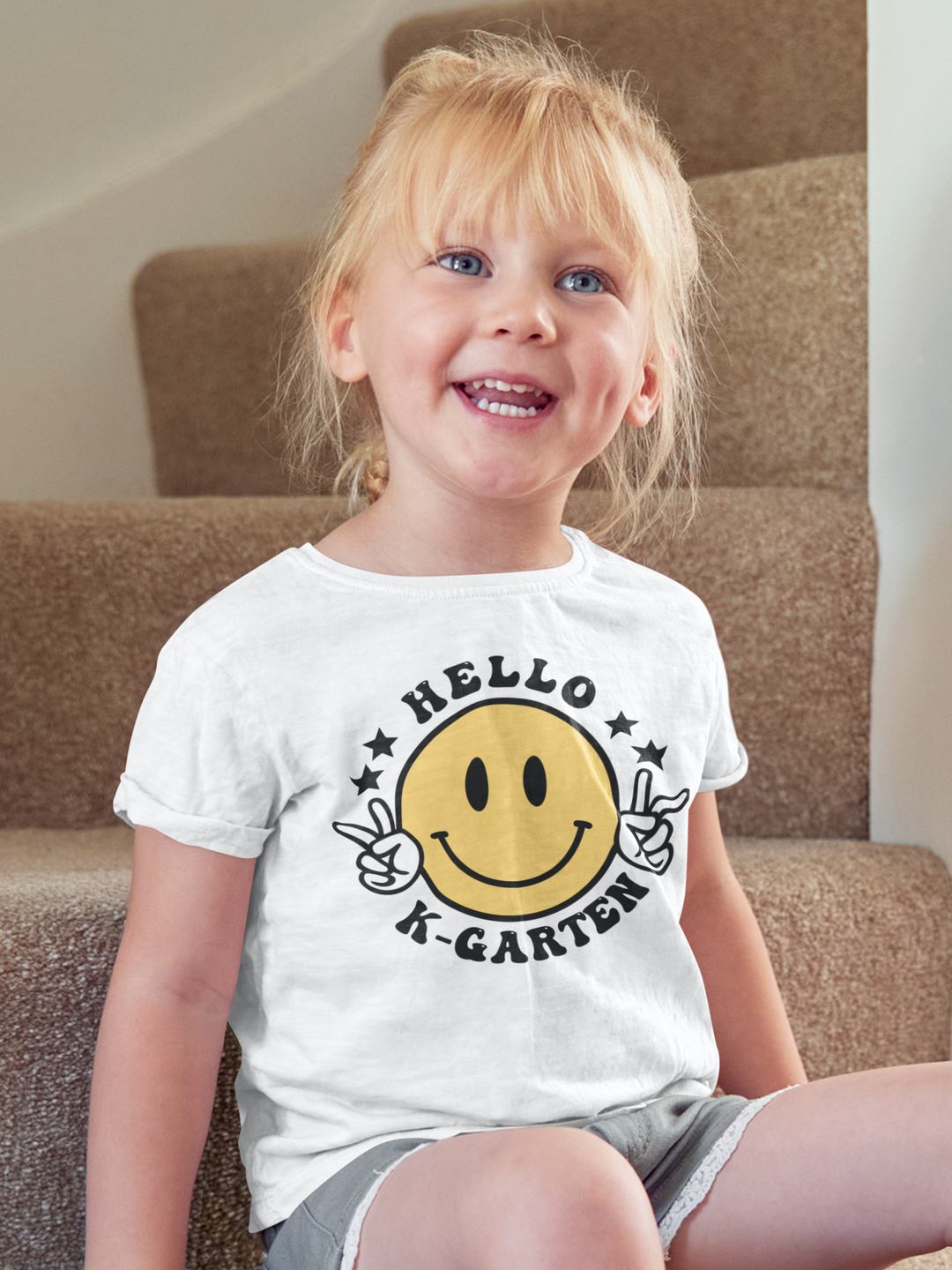 A Hello Kindergarten Toddler Tee featuring a smiling child on stairs, close-ups of eyes, mouth, and nose. Soft 100% cotton, light fabric, tear-away label, perfect for sensitive skin. Ideal for first adventures.