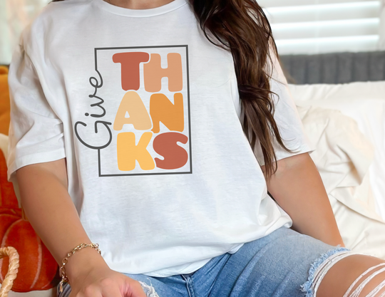 A woman in a white Give Thanks Tee, sitting on a bed. Garment-dyed, 100% ring-spun cotton, relaxed fit, durable double-needle stitching, no side-seams for tubular shape.