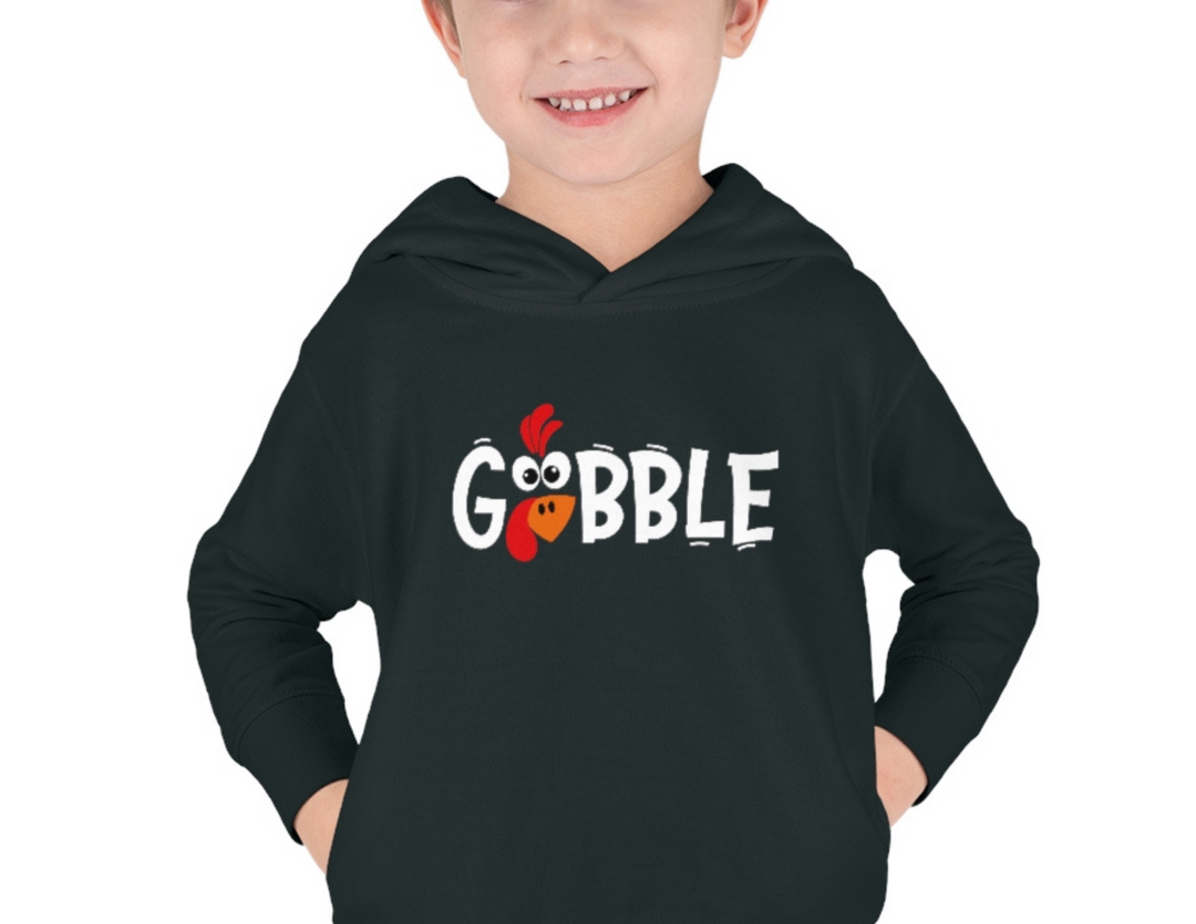 A toddler in a black hoodie with hands in pockets. Gobble Toddler Hoodie: jersey-lined, double-needle hemmed hood, cover-stitched details, side seam pockets for coziness. 60% cotton, 40% polyester. Designed for comfort and durability.