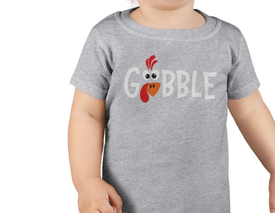 A toddler wearing a grey Gobble Toddler Tee, a soft and durable shirt perfect for sensitive skin. Made of 100% combed ringspun cotton, light fabric, classic fit, tear-away label, and true to size.