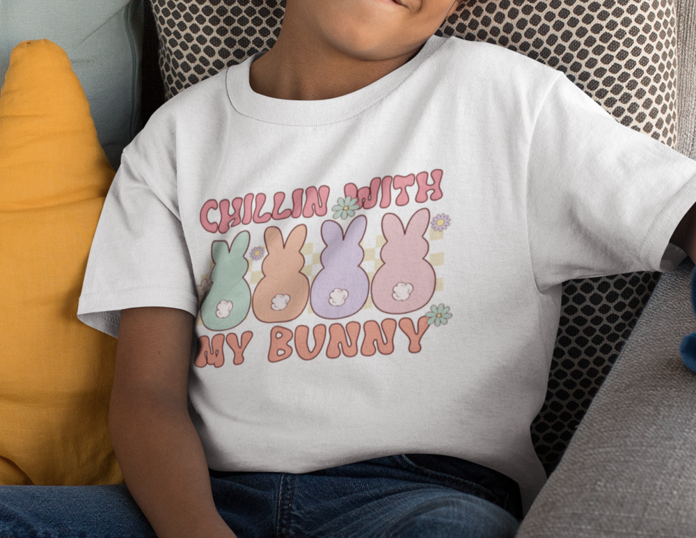 A boy lounging on a couch, wearing the Chillin With My Bunnies Kids Tee. Close-ups of a white shirt, fabric, and adorable bunny details. Perfect for everyday wear, 100% cotton, lightweight, classic fit.