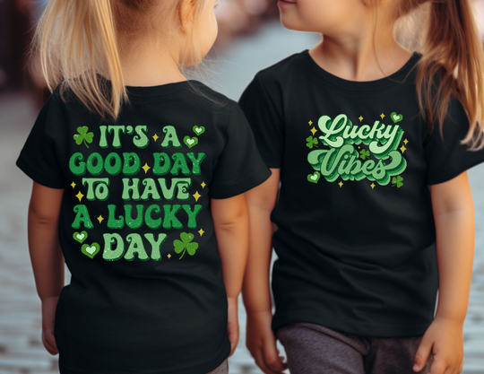 Two girls wearing matching Good Day Lucky Day Toddler Tee, featuring green text and clovers on black shirts, showcasing a casual, playful style.