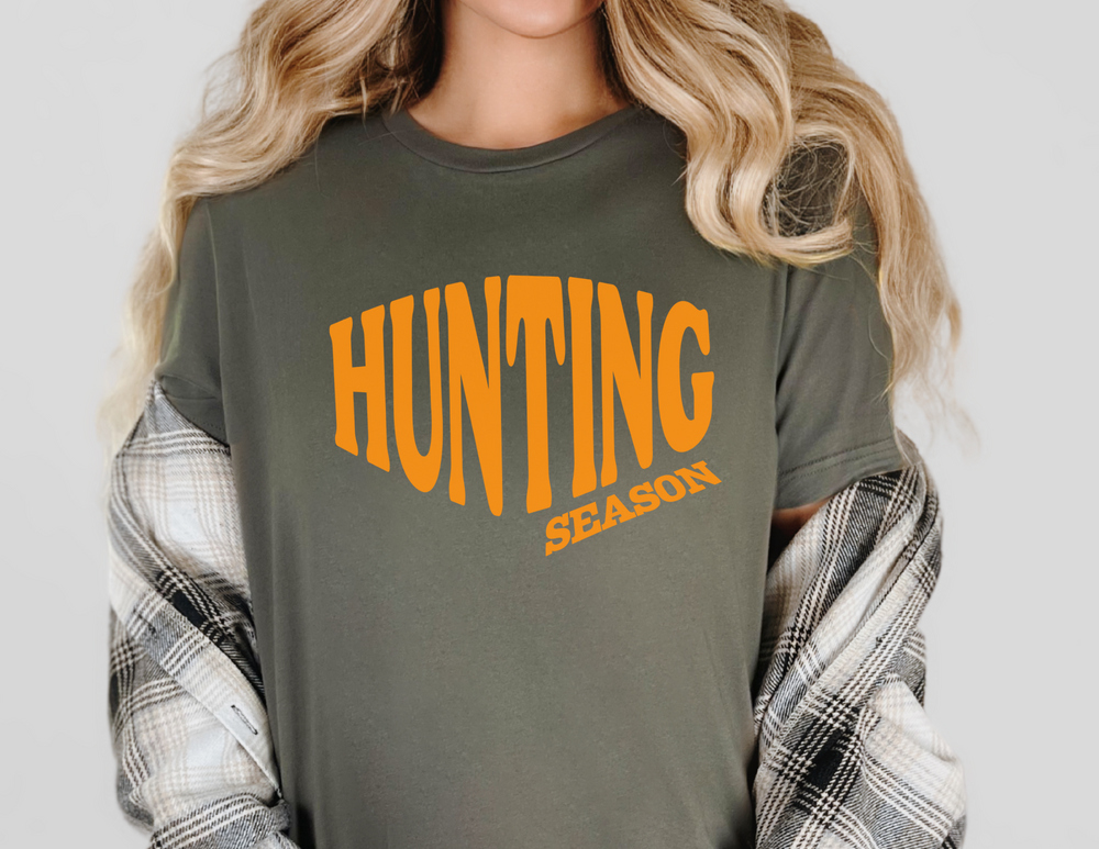 A person wears a muted olive-green HUNTING SEASON tee, slipping off a plaid shirt, showcasing relaxed, durable Comfort Colors 1717 fabric.