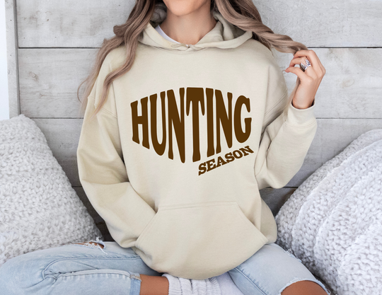 Person in pale beige Hunting Season hoodie, seated cross-legged on a couch, wearing light-wash jeans, surrounded by textured cushions. Perfect for cozy, stylish warmth.