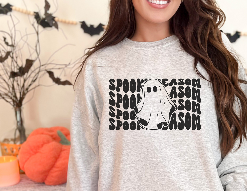 A woman in a grey shirt with a ghost design smiles at the camera. Product: Spooky Season Crewneck. Unisex heavy blend sweatshirt, ribbed knit collar, no itchy seams. Ideal for comfort, made from 50% cotton, 50% polyester.
