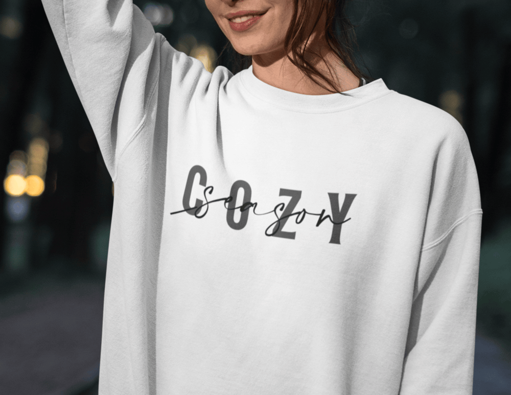 A woman in a white Cozy Season Crewneck sweatshirt, showcasing a logo close-up. Comfortable unisex blend with ribbed collar, no itchy seams. Perfect for casual wear.
