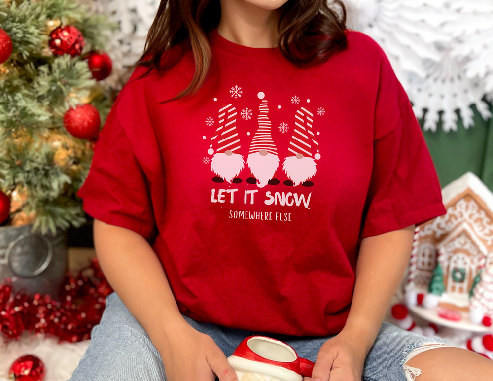 A Let it Snow Somewhere Else Tee featuring a woman in a red shirt, with Christmas-themed elements like gnomes and a gingerbread house. Unisex jersey tee made of soft cotton, with ribbed knit collars and quality print.