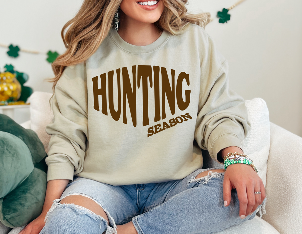Person wearing HUNTING SEASON sweatshirt, paired with ripped jeans and beaded bracelets. St. Patrick’s Day decor in background suggests a festive setting.