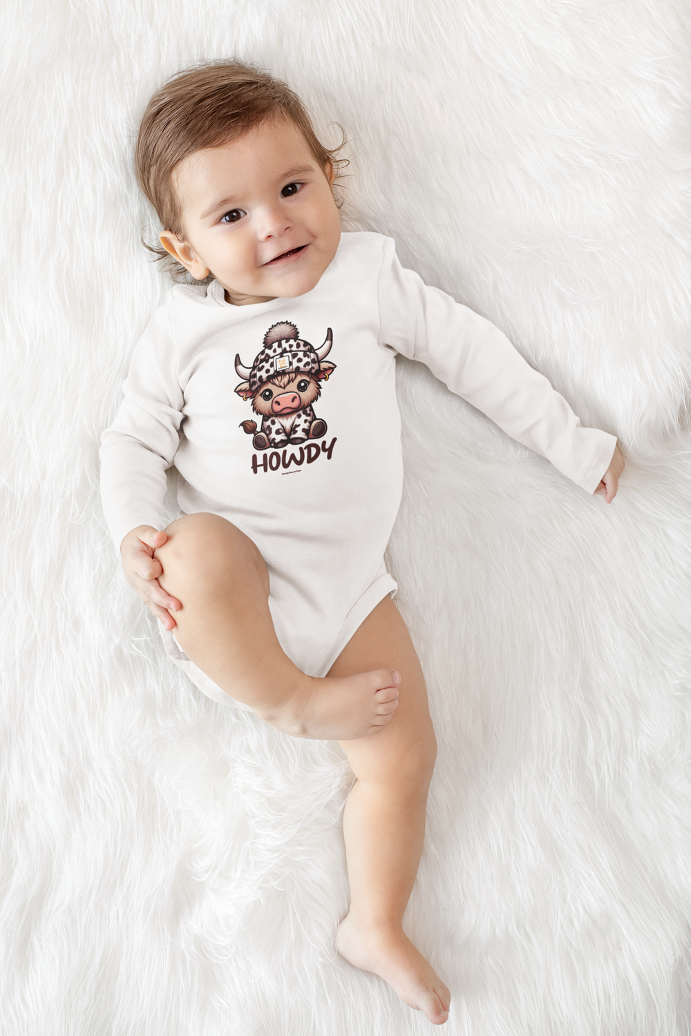 A baby lies on a white blanket, showcasing the Howdy Long Sleeve Onesie for infants. Made of 100% cotton, with ribbed bindings for durability and plastic snaps for easy changing. From Worlds Worst Tees.