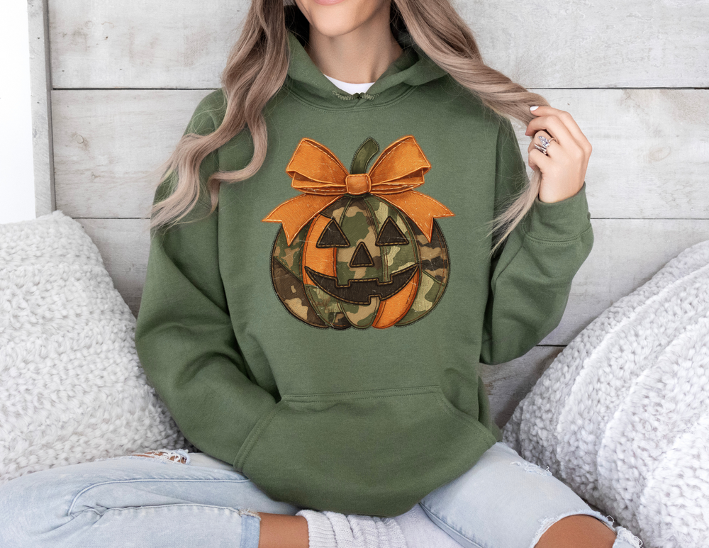 Person seated on couch wearing a forest-green Jackie O Hoodie featuring a camo and orange jack-o-lantern graphic with an orange bow, paired with distressed jeans.