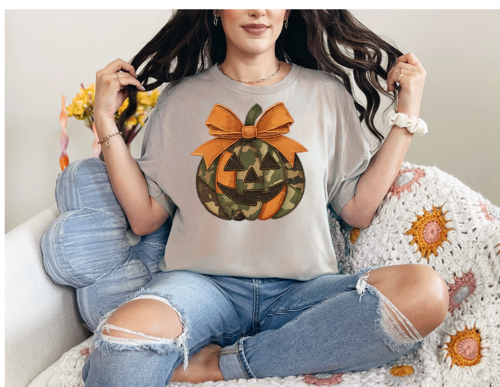 Woman in ripped jeans and light-gray Jackie O Tee with a split orange and camouflage jack-o-lantern graphic, sitting on a colorful crochet blanket.