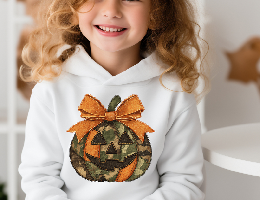 Jackie O Youth Hoodie features a camouflage and orange jack-o'-lantern with a bow, worn by a smiling girl, showcasing its comfortable fit and stylish design.