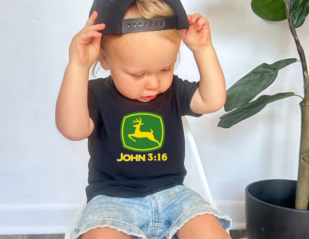 Toddler wearing John 3:16 Baby Tee sits on a white chair, sporting a backward black cap, with light denim shorts next to a potted plant.