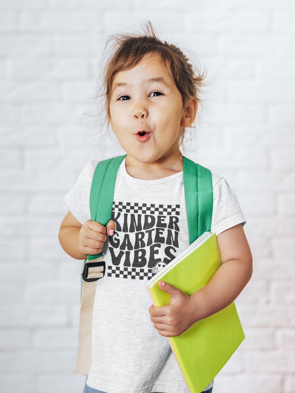 A Kindergarten Vibes Toddler Tee featuring a child with a backpack and a book. Soft 100% combed ringspun cotton, light fabric, tear-away label, and a classic fit for comfort and durability. Ideal for young explorers.