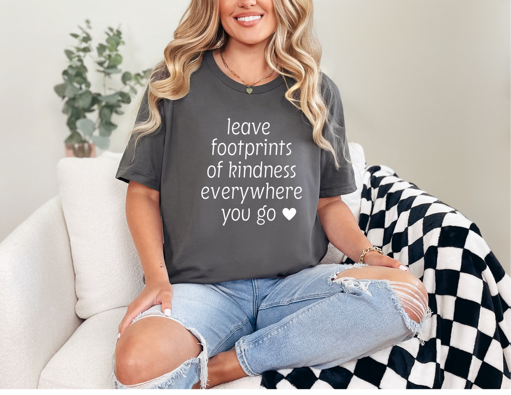 Woman in a Leave Kindness Tee, smiling on a sofa, wearing light-wash jeans, highlighting the tee's relaxed fit and comfortable style.