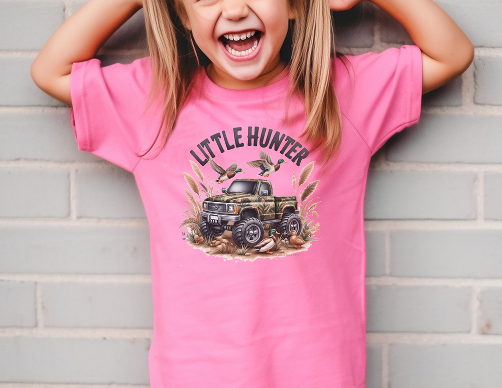 A Little Hunter Kids Tee in pink, featuring a girl in a pink shirt, smiling. Made of 100% combed ringspun cotton for comfort and agility. Perfect for studying or playtime.