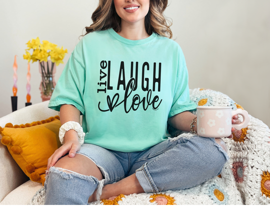 A woman sits on a couch holding a mug, embodying comfort and relaxation with the Live Laugh Love Tee. Made of 100% ring-spun cotton, this cozy tee features a relaxed fit and durable double-needle stitching.
